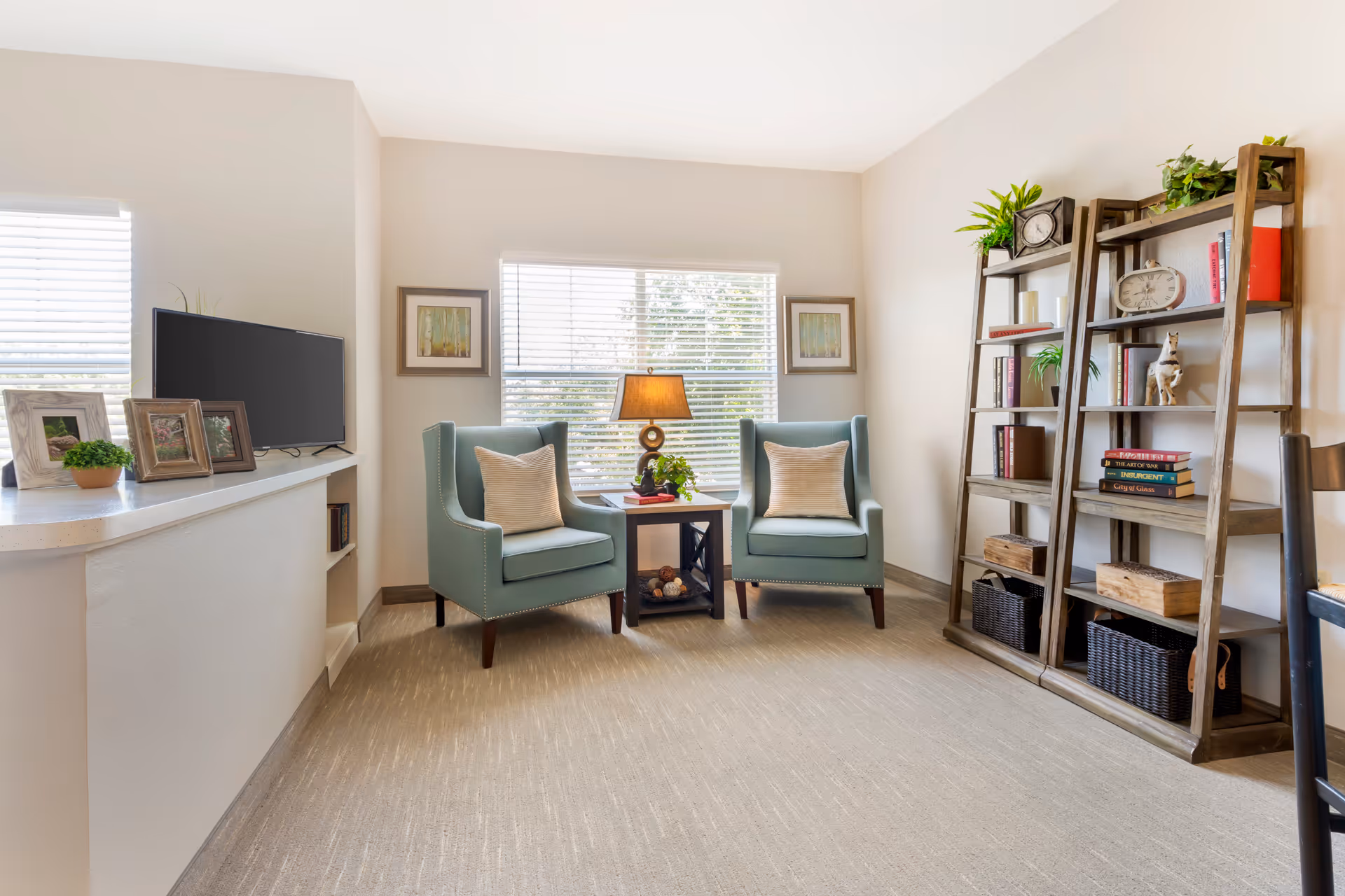 A cozy living room area with two teal armchairs with beige pillows facing a small wooden side table with a lamp and a plant. Behind the chairs is a window with blinds and two framed pictures on either side. To the right, there are two wooden ladder-style bookshelves with books, plants, and decorative items. On the left, a TV sits on a white counter with picture frames and a small potted plant.