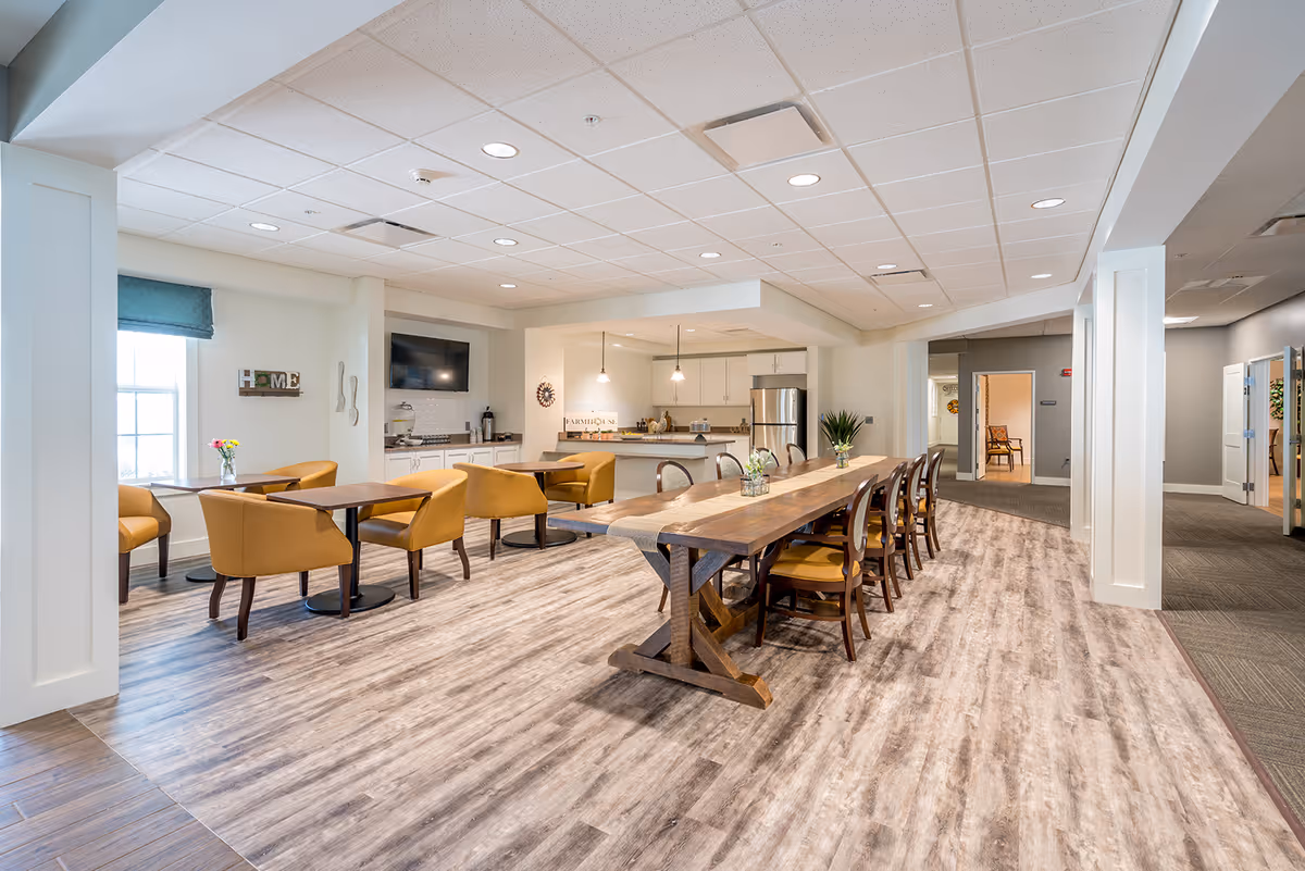 A spacious and well-lit dining area in a senior living facility featuring a long wooden table with chairs on one side and smaller tables with mustard yellow upholstered chairs near a window. The room has wood-patterned flooring, a kitchen area with stainless steel appliances, pendant lighting, and a wall-mounted TV. Decorative elements include a 'HOME' sign and fresh flowers on the tables.
