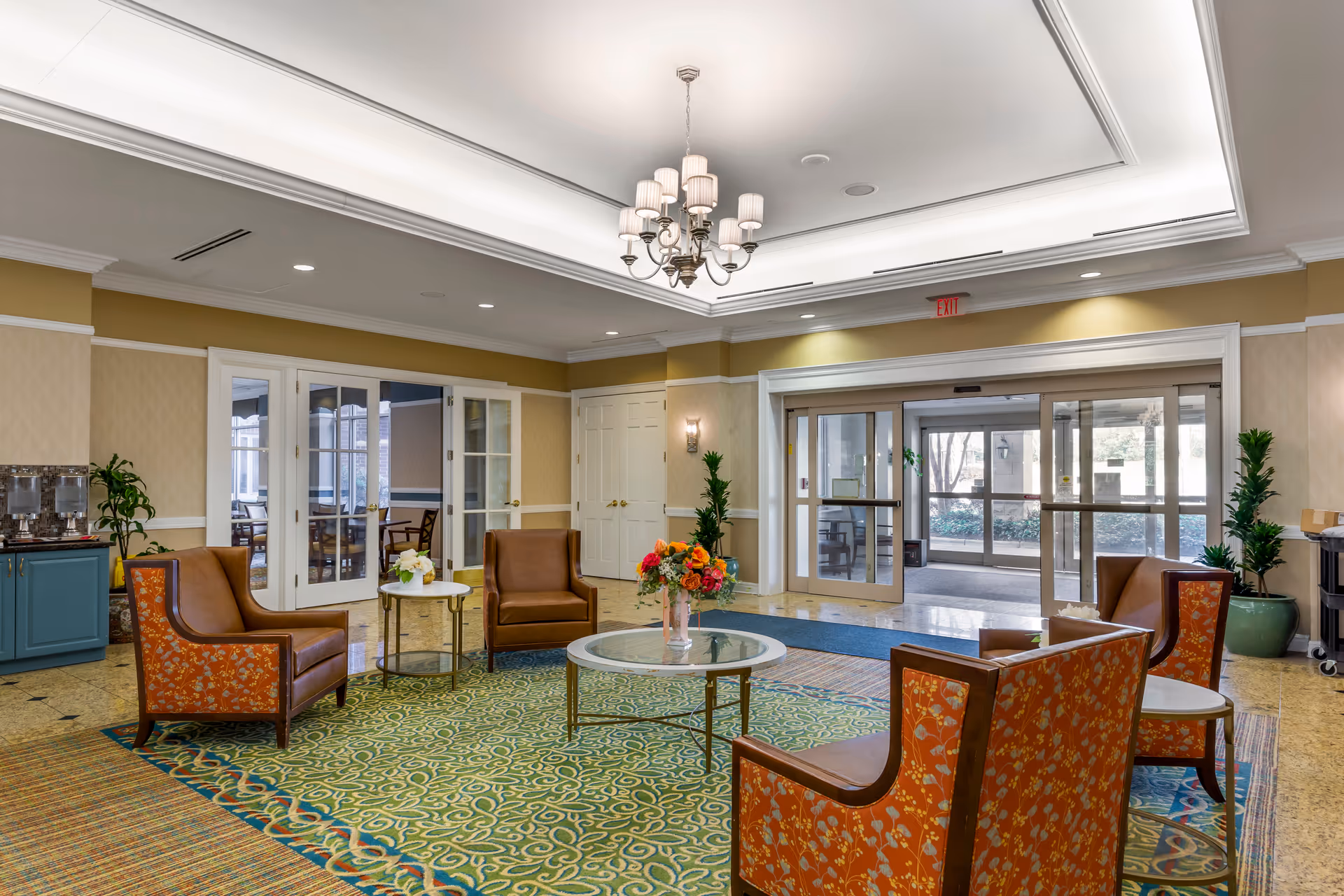 Bright lobby with upholstered armchairs arranged around a central glass coffee table, a floral centerpiece, and automatic glass entrance doors.