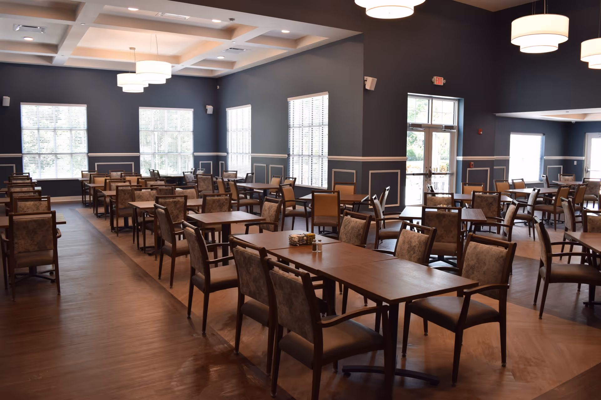 A spacious dining room with multiple wooden tables and cushioned chairs arranged neatly. The room has large windows with blinds allowing natural light to enter, dark blue walls with white trim, and modern ceiling lights hanging from a coffered ceiling. There are exit doors visible in the background.