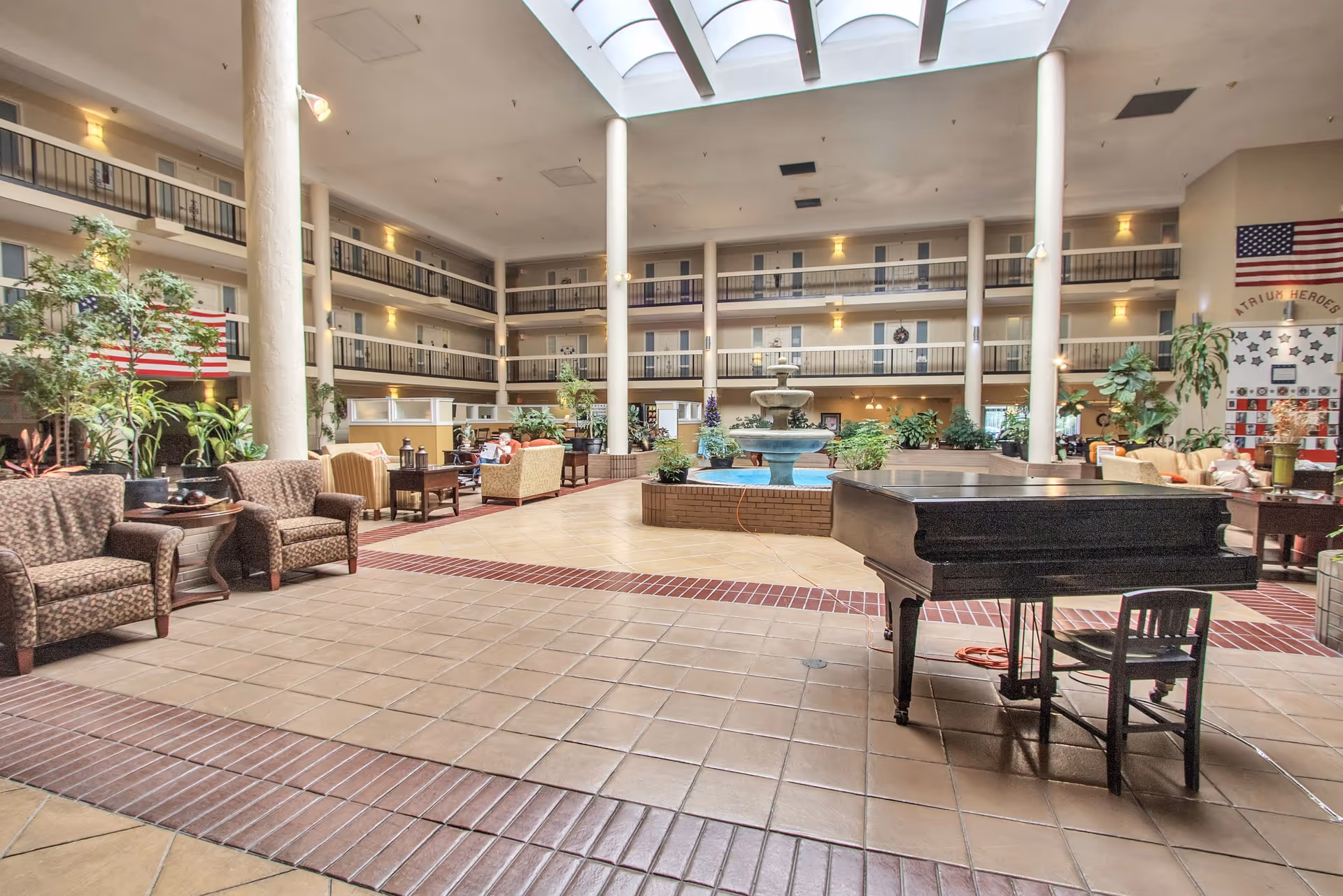 Spacious indoor atrium area with multiple seating arrangements including armchairs and sofas, a large fountain in the center, a black grand piano with a chair, several potted plants, and a three-story balcony with doors overlooking the atrium. An American flag and a wall display labeled 'ATRIUM HEROES' are visible on the far wall.