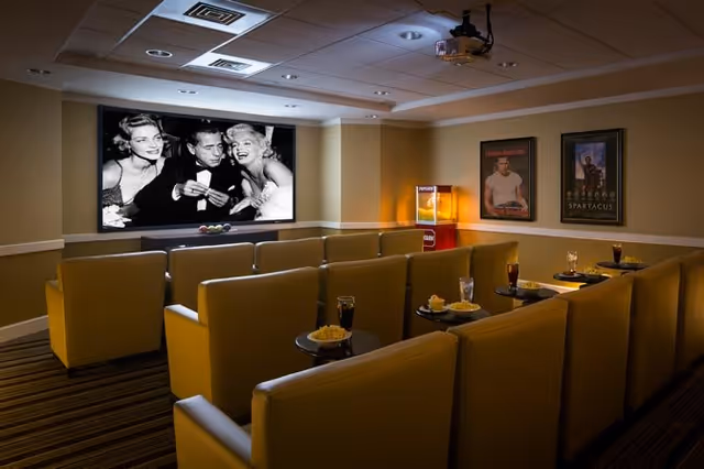 A small movie theater room with rows of yellow cushioned seats and small round tables holding popcorn and drinks. A black and white movie is playing on a large screen at the front. The room has a popcorn machine and framed movie posters on the walls.