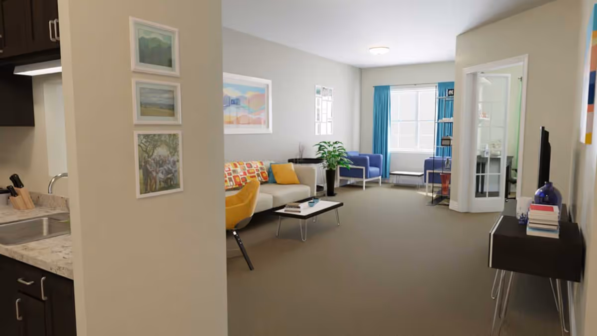 A bright and spacious living room with a beige sofa adorned with colorful cushions, a yellow chair, two blue armchairs near a window with blue curtains, a coffee table with books and a cup, a potted plant, and framed artwork on the walls. To the left, part of a kitchen with a sink and countertop is visible, and to the right, a black console table with books and decorative items is against the wall.