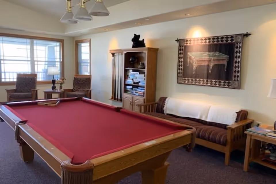 A cozy game room with a red felt pool table in the center. Behind the pool table, there is a wooden bookshelf filled with books and a rack holding pool cues. To the right, there is a wooden couch with striped cushions and a white throw blanket. On the wall above the couch, a framed tapestry depicting a pool table is displayed. Two armchairs and a small table with a lamp and flowers are positioned near a large window letting in natural light.