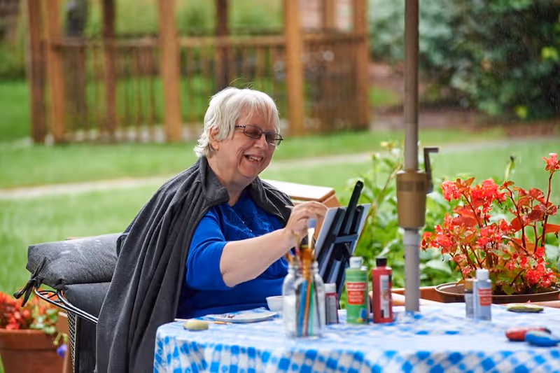 An elderly woman with white hair and glasses is sitting outdoors at a table covered with a blue and white patterned tablecloth. She is smiling and painting on a small canvas set on an easel. Various paint bottles and brushes are on the table. Behind her is a green lawn and wooden garden structures, with red flowers nearby.