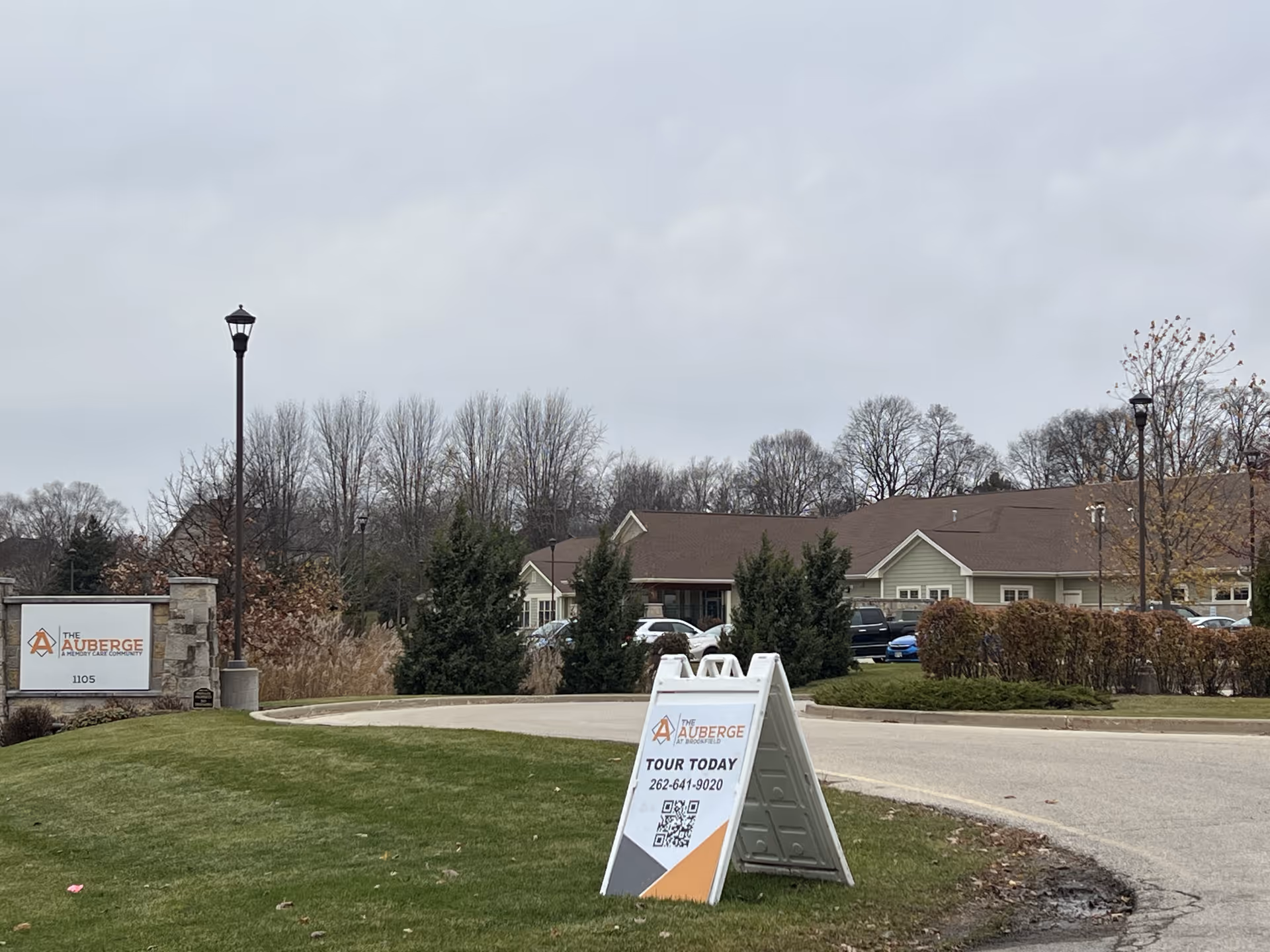 Exterior view of The Auberge at Brookfield facility with a sign displaying the facility name and address, a sandwich board sign inviting people to tour today with a phone number and QR code, a driveway, some parked cars, trees without leaves, and a cloudy sky.