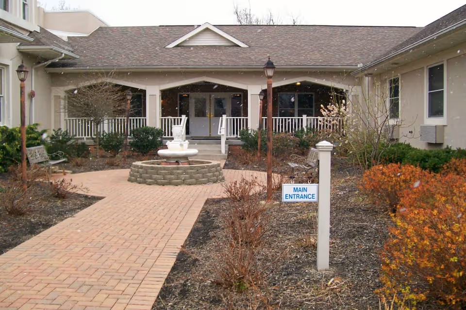 Front courtyard with a brick walkway leading to a covered main entrance, a small fountain, benches, and shrubs.