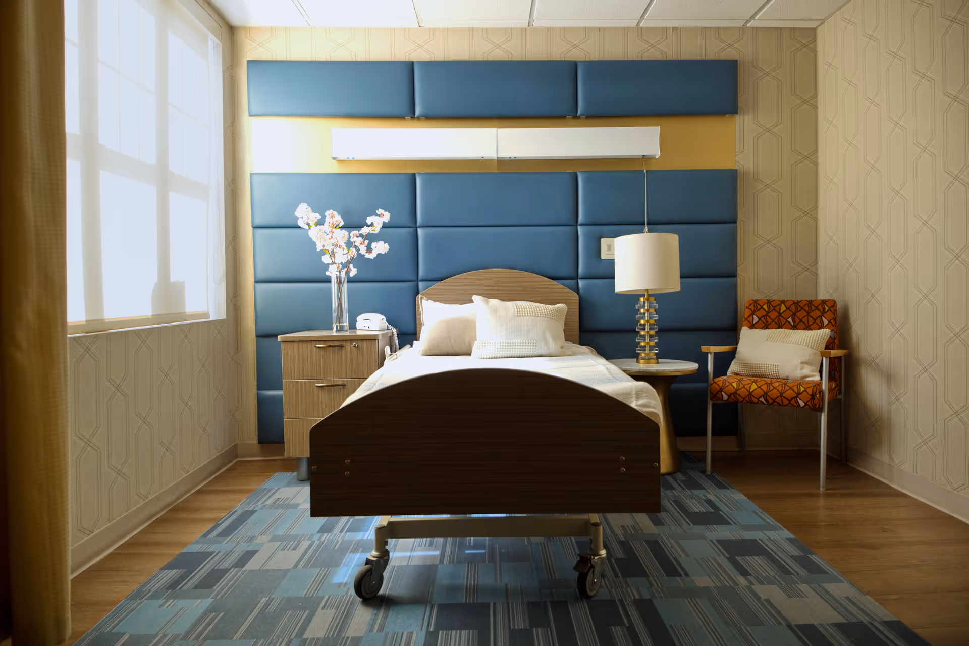 A single bed with white bedding and pillows in a room with a blue padded headboard wall. Next to the bed is a wooden nightstand with a vase of flowers and a telephone. On the other side, there is a round table with a lamp and an orange patterned armchair with a cushion. The room has a large window with sheer curtains and patterned wallpaper.