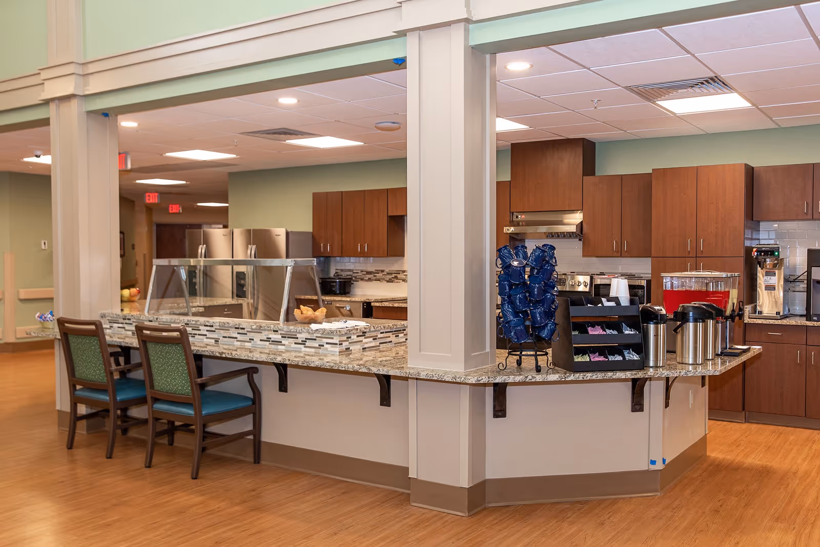 Interior view of a kitchen area in a senior living facility with wooden cabinets, stainless steel appliances, a granite countertop with a serving area, chairs, and beverage dispensers.