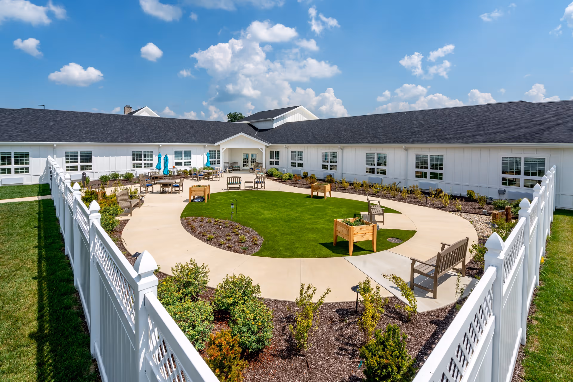Outdoor courtyard area at Sugar Fork Crossing featuring a circular walkway surrounding a grassy lawn with raised garden beds, benches, and patio tables with umbrellas. The courtyard is enclosed by a white fence and surrounded by a single-story white building under a blue sky with scattered clouds.