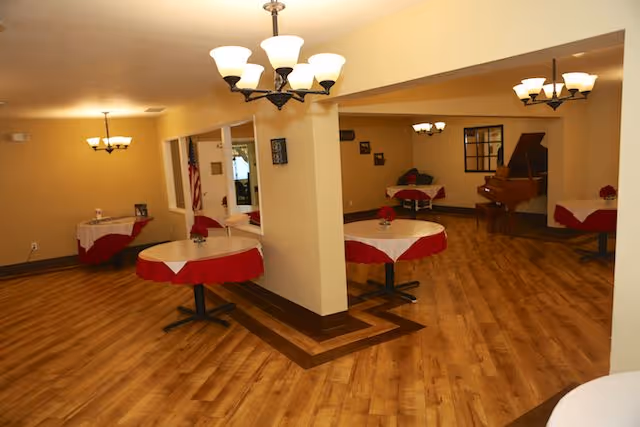 Interior view of a dining area with round tables covered with white and red tablecloths, wooden flooring, and multiple ceiling light fixtures. A grand piano is visible in the background along with some framed pictures on the walls.