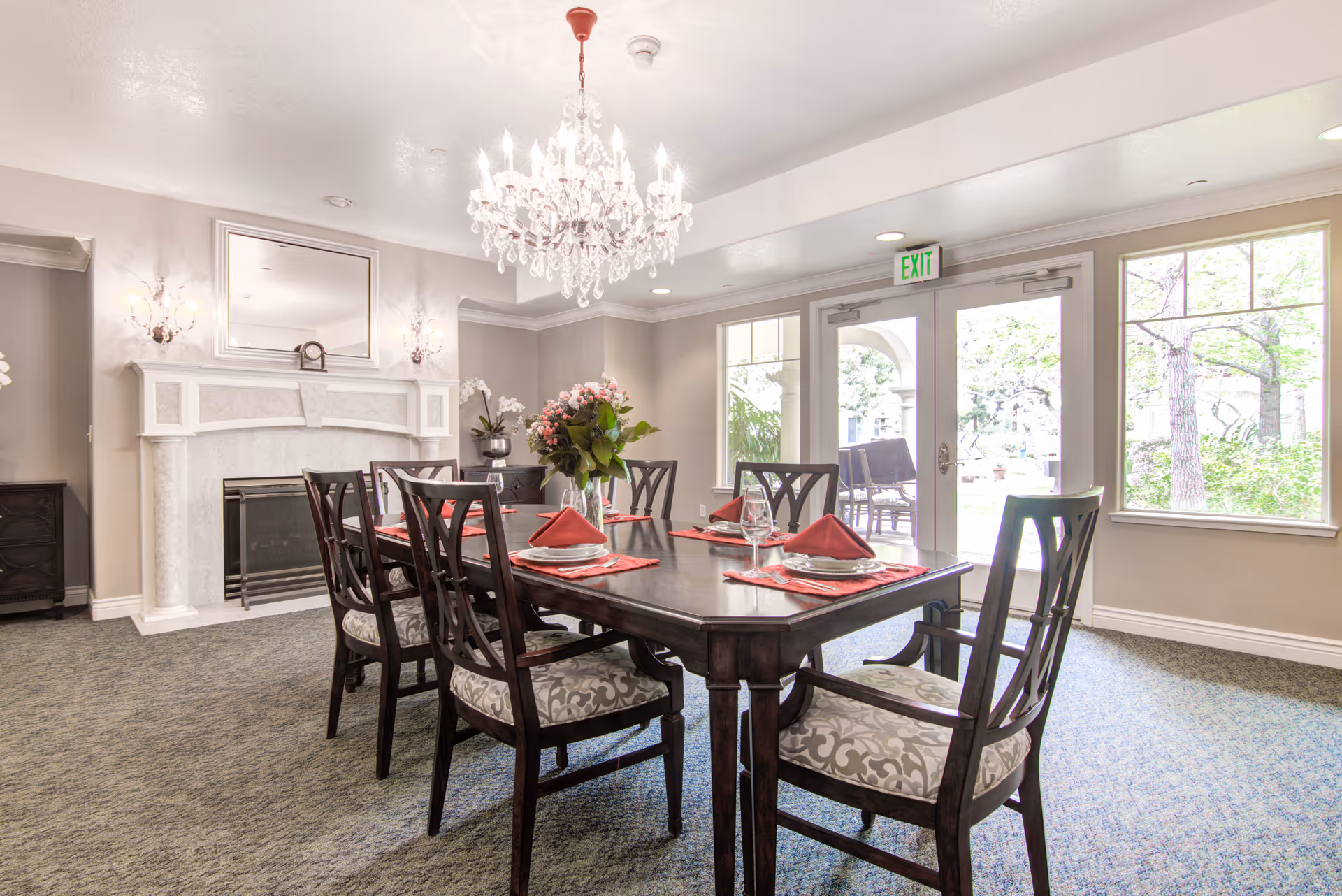 A bright dining room with a dark wooden table set for six people with red napkins and glassware. The room features a white fireplace with a mirror above it, a chandelier hanging from the ceiling, large windows, and a glass door leading outside to a garden area.