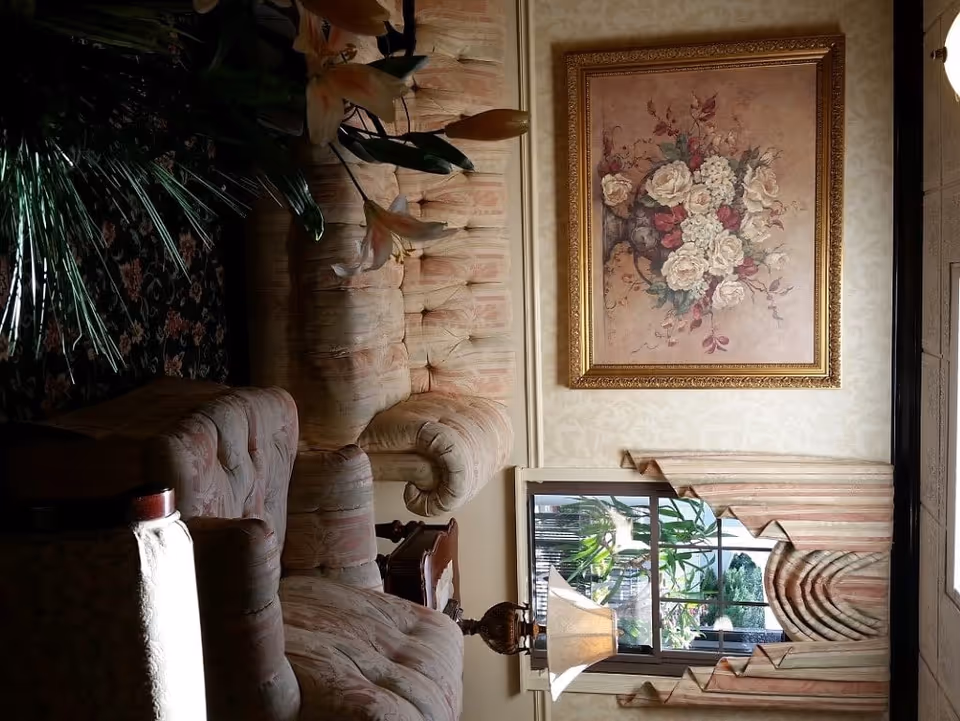 A cozy living room area with a floral-patterned upholstered sofa and armchair. There is a side table with a decorative lamp, a large framed painting of a floral bouquet on the wall, and a window with patterned curtains partially open showing greenery outside.