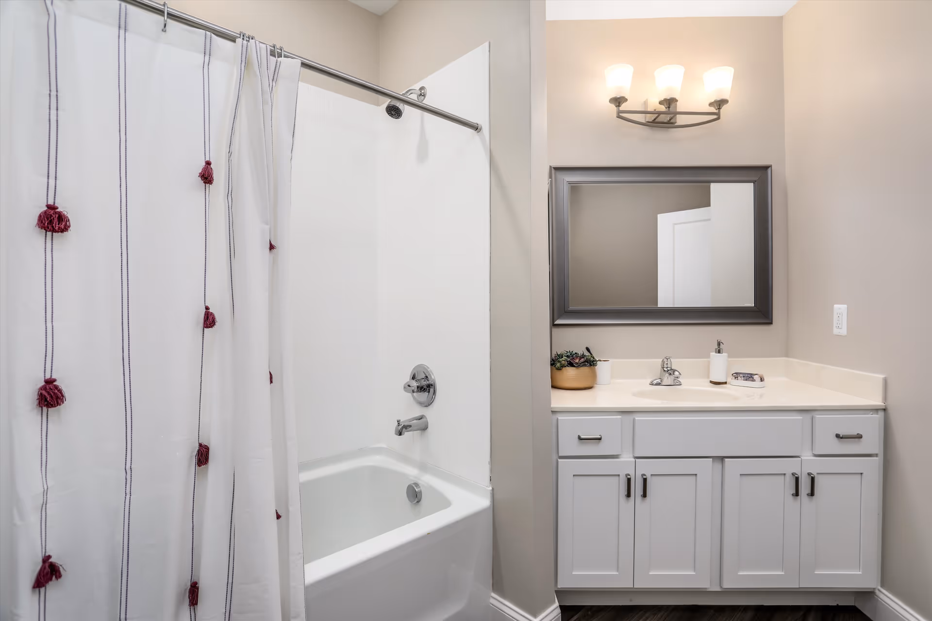 A clean and modern bathroom featuring a white bathtub with a shower curtain decorated with vertical stripes and red tassels. Next to the bathtub is a white vanity with a sink, a large rectangular mirror with a silver frame, and a three-light wall fixture above. The vanity countertop holds a small plant, a soap dispenser, and a soap dish.