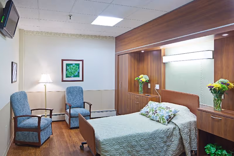 A neatly arranged senior living bedroom with a single bed, floral pillows, wooden cabinetry, two upholstered chairs and a floor lamp.