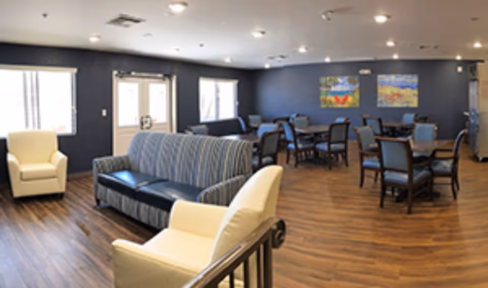 A spacious common area with wooden flooring, featuring a striped blue sofa, two cream armchairs, and several tables with chairs arranged for dining or socializing. The walls are painted dark blue and decorated with colorful paintings. Large windows and a glass door allow natural light to enter the room.