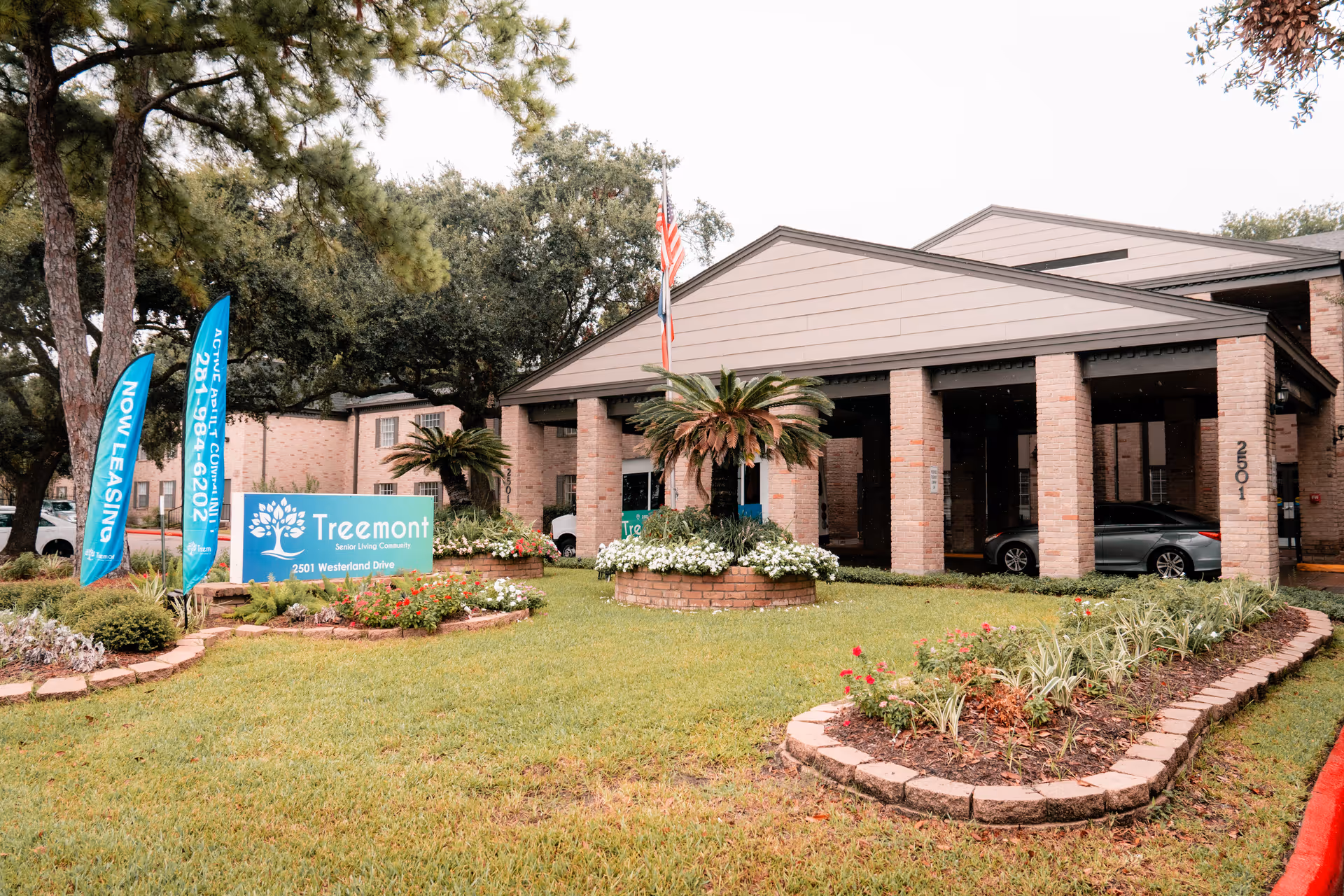 Front entrance of Treemont Senior Living with a sign, flags, landscaped lawn, and a covered drive-through entrance.