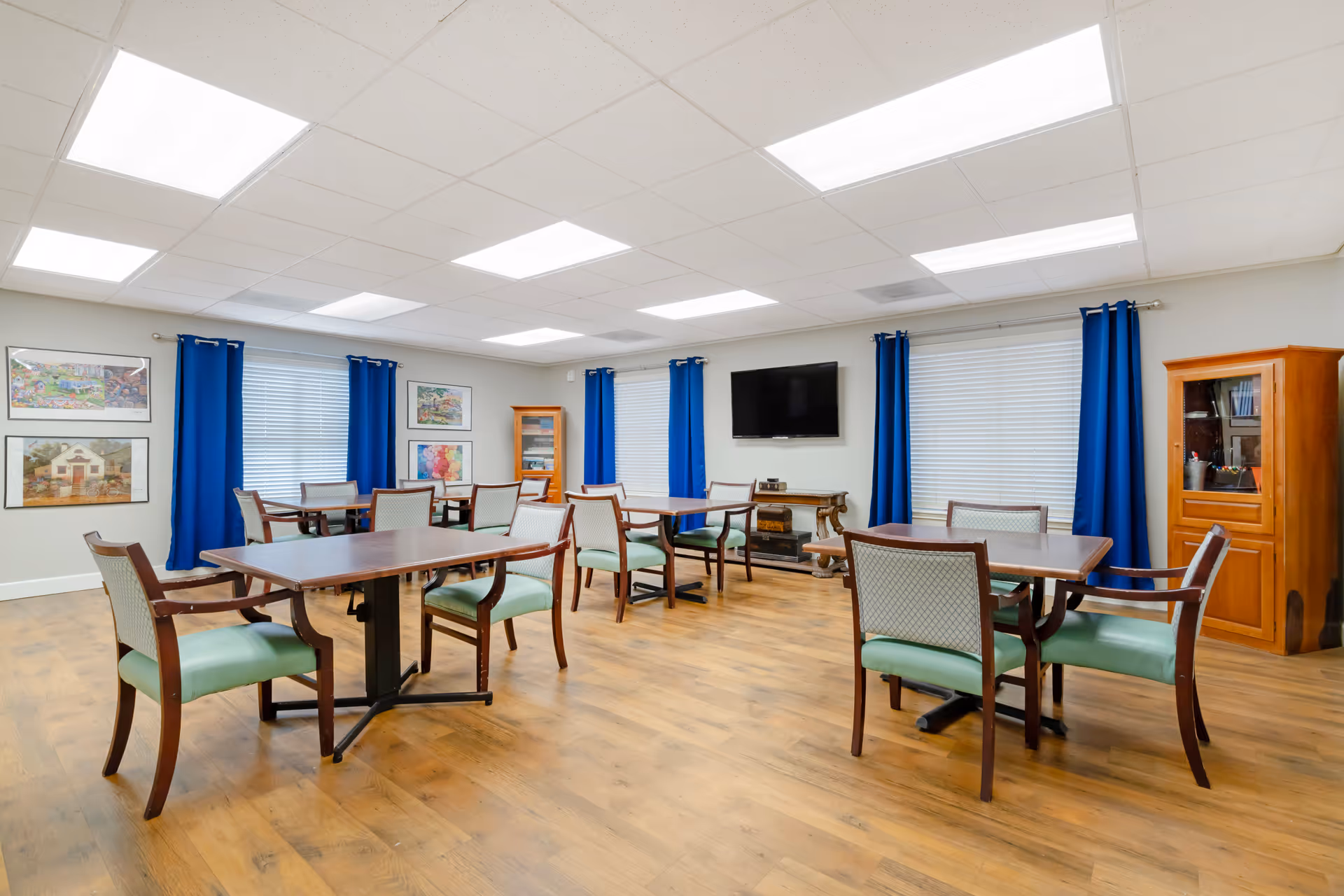 A bright and spacious dining room with wooden floors, several tables and chairs with green cushions, blue curtains on the windows, framed artwork on the walls, a flat-screen TV mounted on the wall, and wooden cabinets.