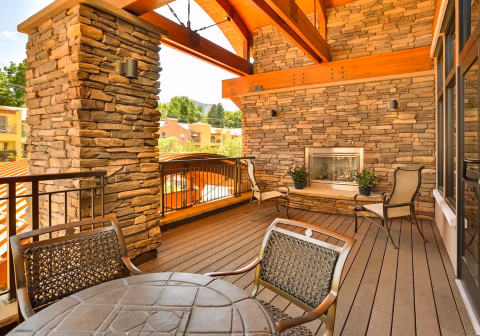 Covered outdoor patio with stone pillars and a wood-beamed ceiling, featuring a fireplace, table and chairs overlooking neighboring buildings.