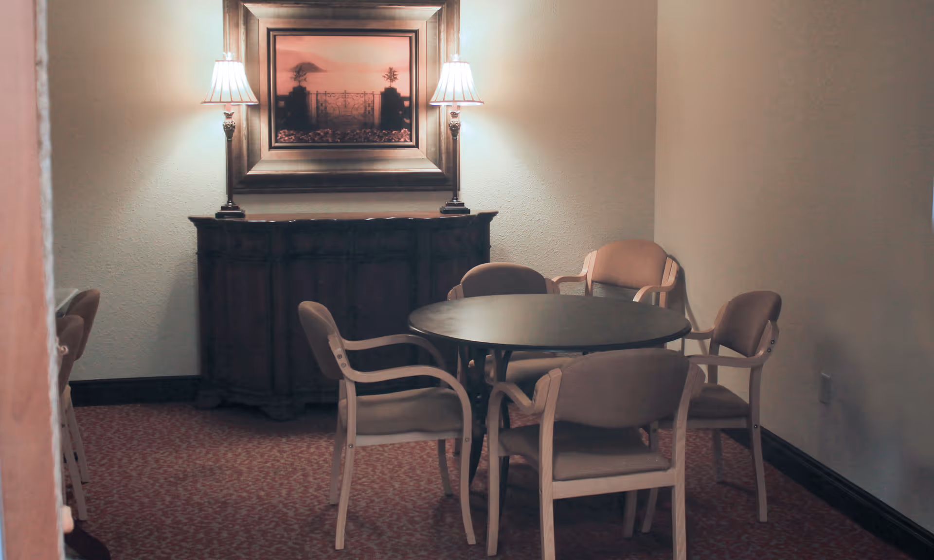 Small dining room with a round table surrounded by five upholstered chairs, a wooden sideboard with two lamps, and a framed painting on the wall.