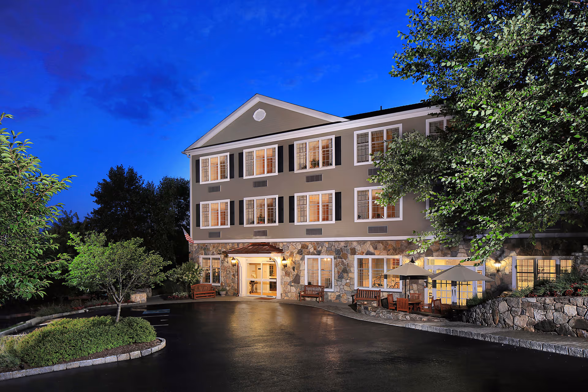 Exterior view of a three-story senior living facility building at dusk with lit windows, stone facade on the lower level, benches, umbrellas, and surrounding trees and landscaping.