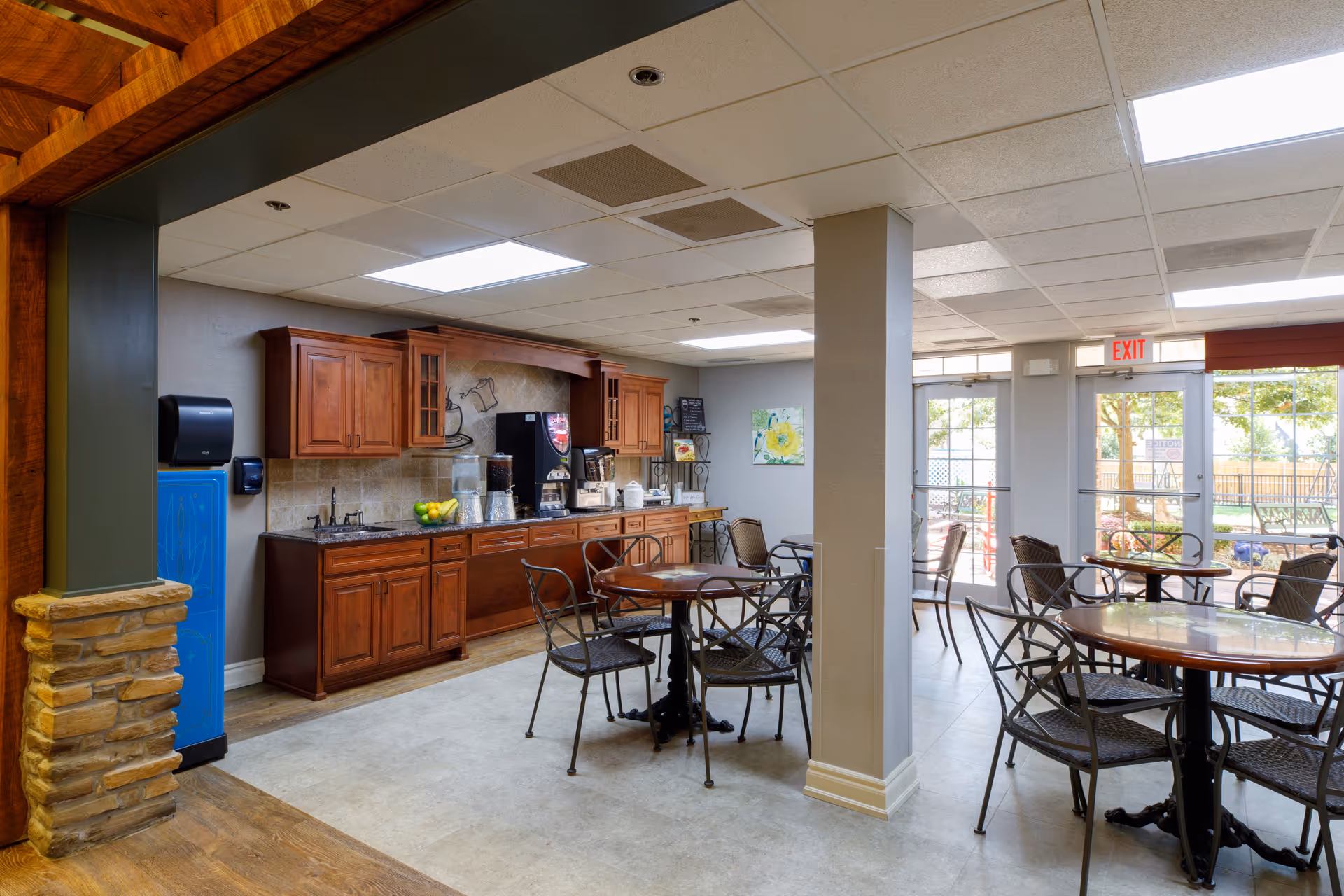 A bright dining area with round tables and metal chairs, a kitchenette with wooden cabinets, a coffee machine, and a water dispenser. Large windows and glass doors let in natural light and provide a view of an outdoor garden area.