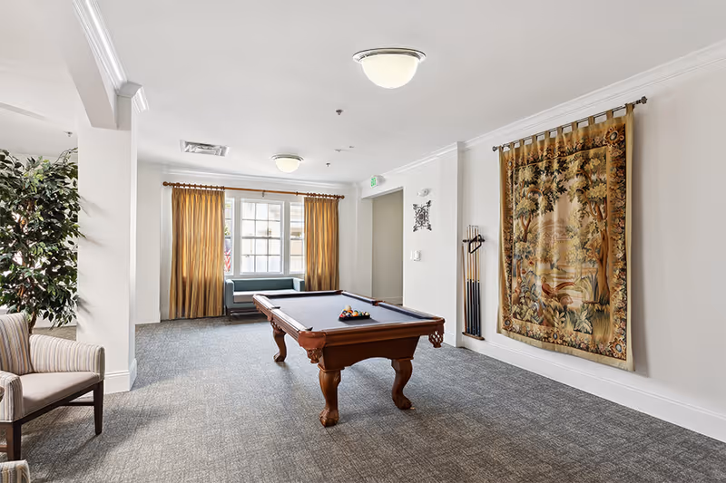 Bright communal game room with a pool table, seating, a large window with curtains, and a decorative tapestry on the wall.