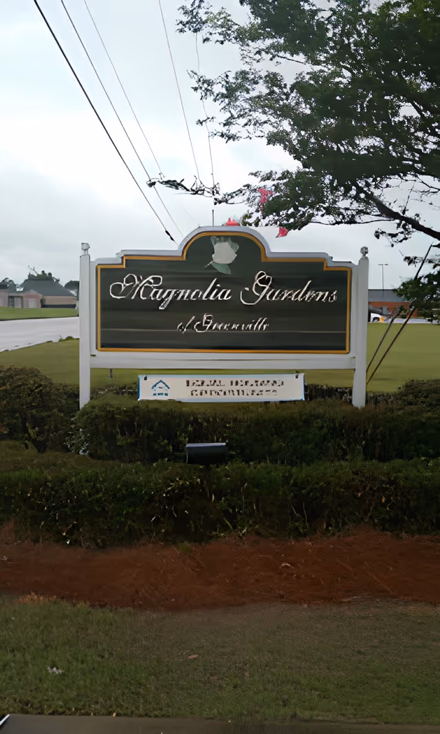 Outdoor view of a green and white sign for Magnolia Gardens of Greenville, surrounded by bushes and trees, with a cloudy sky in the background.