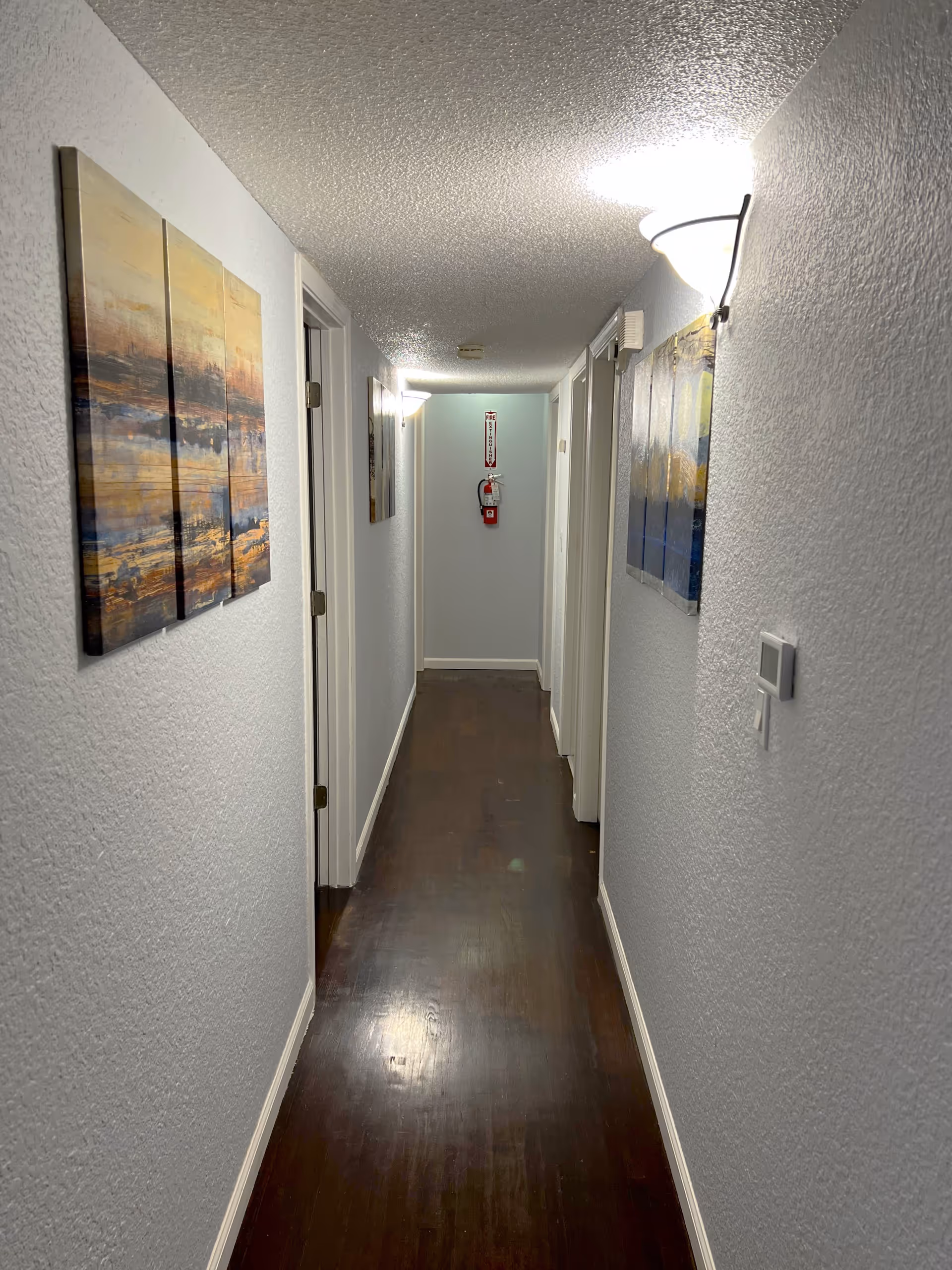 A narrow hallway with dark wooden flooring and light gray textured walls. The hallway is illuminated by wall-mounted lights on both sides. There are several doors along the hallway, and two abstract paintings hang on the walls. A fire extinguisher is mounted on the far wall beneath a fire safety sign.