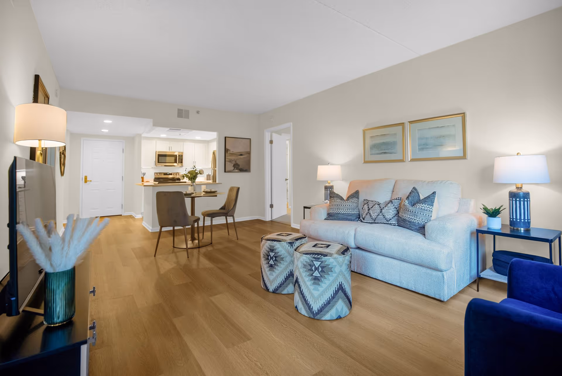 A bright and modern living room with a light-colored sofa adorned with patterned cushions, two patterned ottomans, a blue armchair, and two side tables with lamps and a small plant. The room has wooden flooring and light-colored walls with two framed artworks above the sofa. In the background, there is a small dining area with a round table and two chairs, and a kitchen with white cabinets and stainless steel appliances. A TV is visible on the left side of the image, placed on a black stand with a decorative vase containing pampas grass.