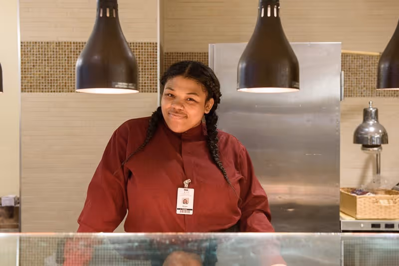 A woman with braided hair wearing a maroon uniform and an ID badge stands behind a glass counter in a kitchen or food service area with hanging black pendant lights above her.