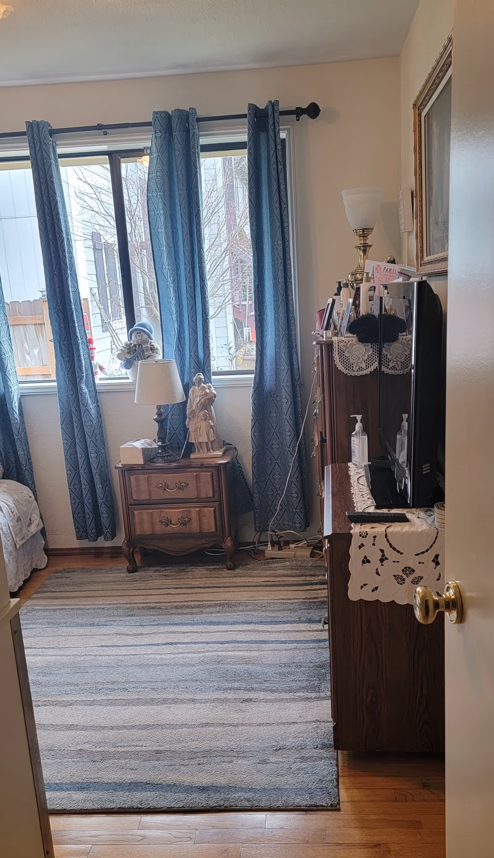 A cozy bedroom with a wooden nightstand holding a lamp, tissue box, and a figurine. Blue patterned curtains hang over a window showing an outdoor view. A wooden dresser with a TV on top is on the right side, decorated with a lace runner and various items. A striped area rug covers the wooden floor.