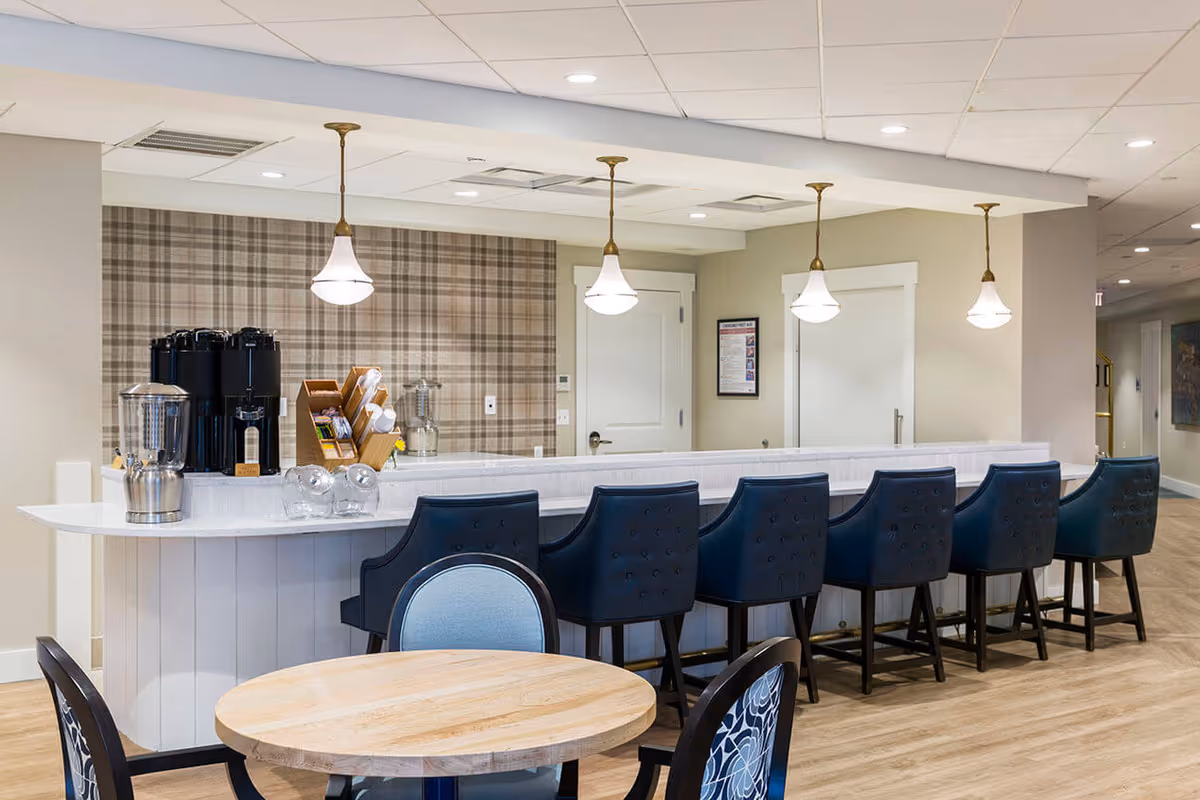 Interior view of a senior living facility's dining or common area featuring a long white counter with six dark blue cushioned bar stools. Above the counter, there are four hanging pendant lights with white shades. On the counter, there are beverage dispensers, a wooden organizer with cups, and some glass containers. In the foreground, there is a round wooden table with two chairs, one with a light blue cushion and the other with a patterned backrest. The walls are light-colored with a plaid accent wall behind the counter, and the floor is light wood.