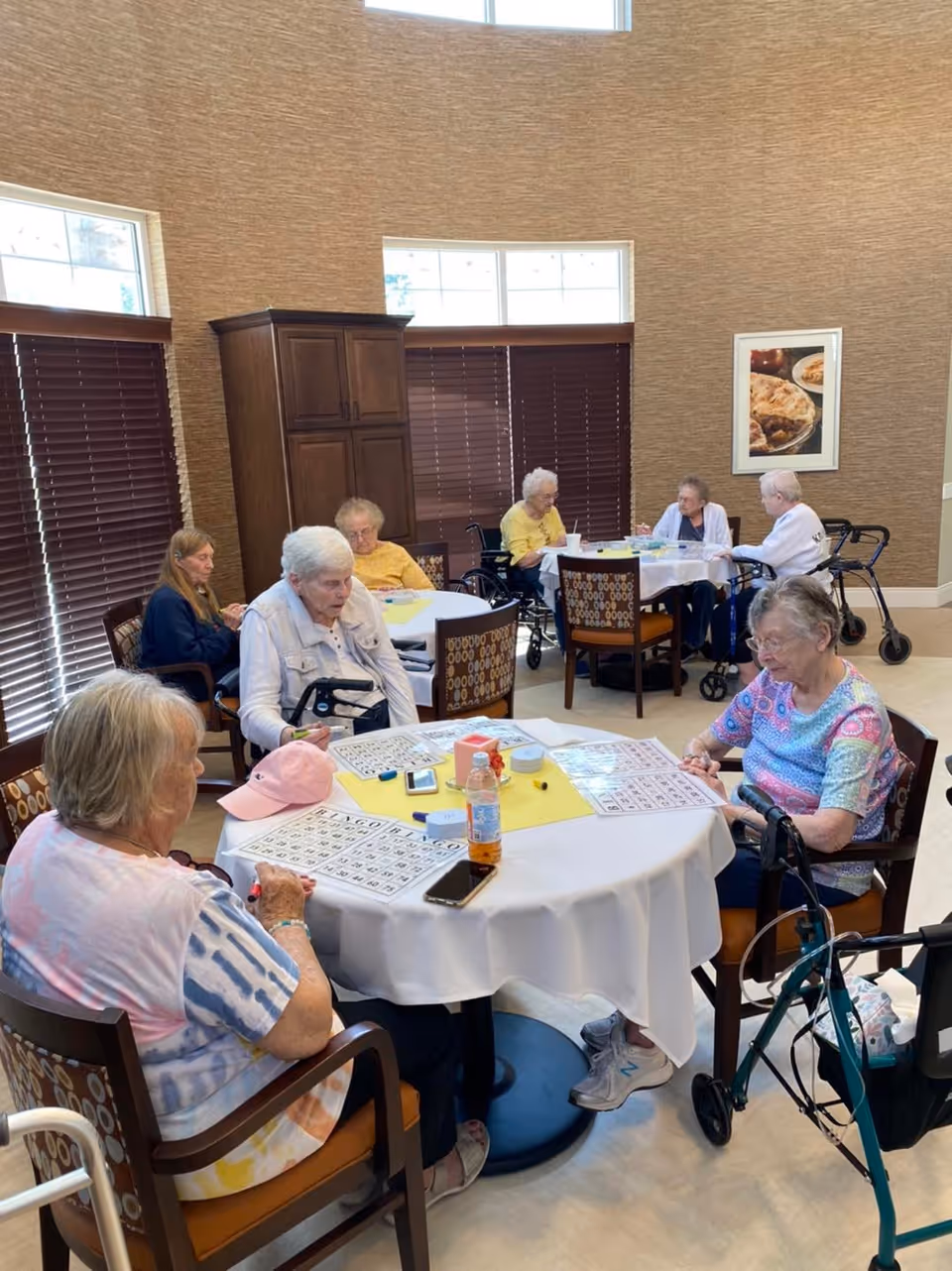 A group of elderly women sitting around two round tables in a well-lit room with large windows and brown blinds. They appear to be playing bingo, with bingo cards, markers, and a bottle of drink on the tables. Some women are using walkers and wheelchairs. The room has beige walls, a wooden cabinet, and a framed picture of food on the wall.