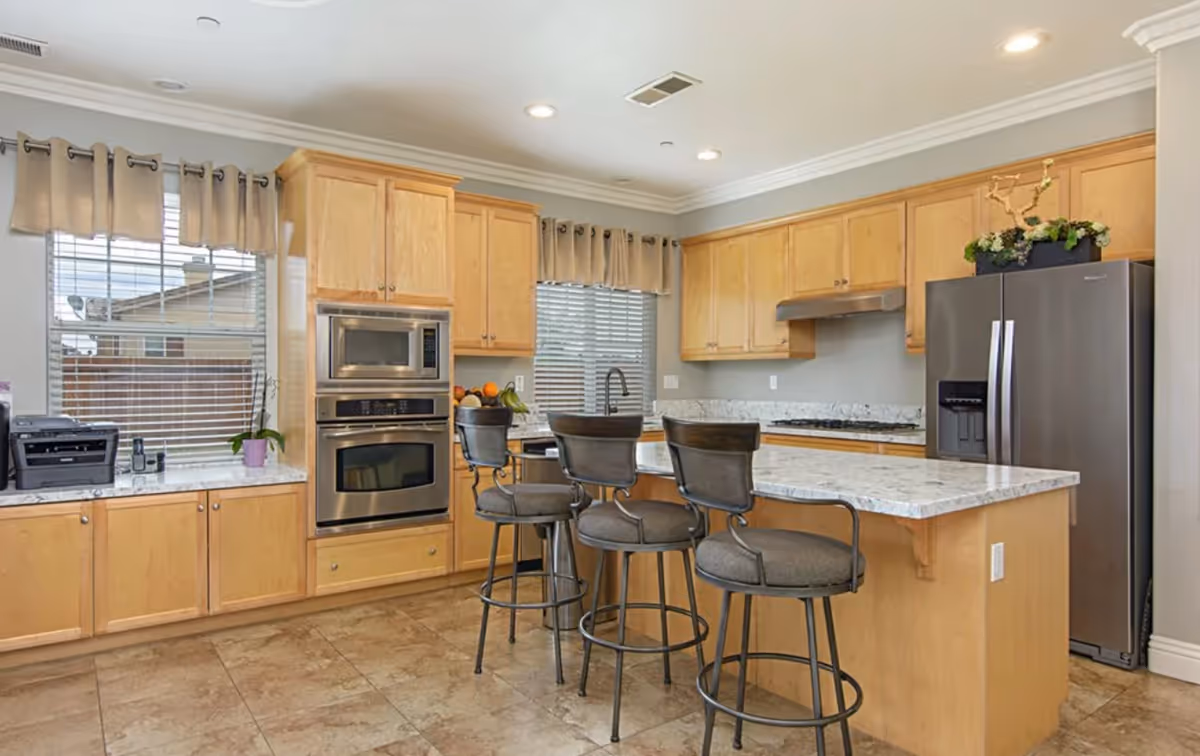 A modern kitchen with light wood cabinets, a marble countertop island with three cushioned bar stools, stainless steel appliances including a refrigerator, oven, and microwave, and two windows with beige curtains.