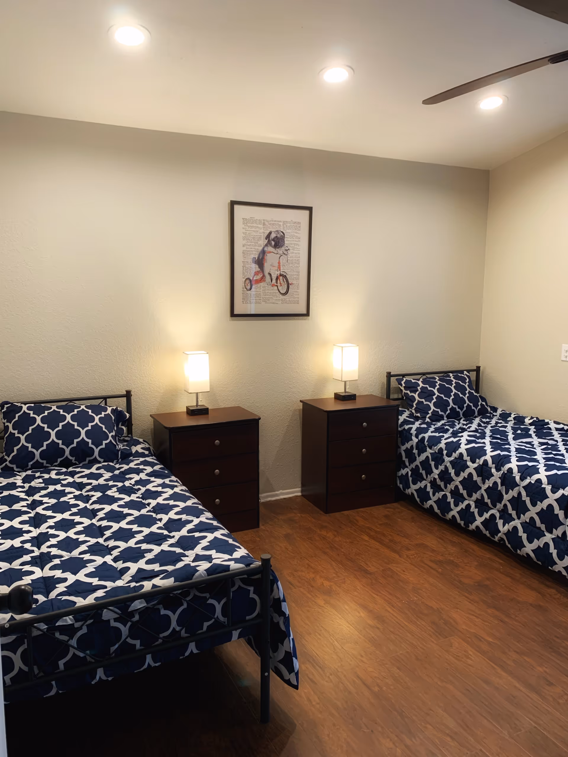 A tidy bedroom with two twin beds dressed in navy patterned bedding, two dark wood nightstands with lamps, and framed artwork on the wall.