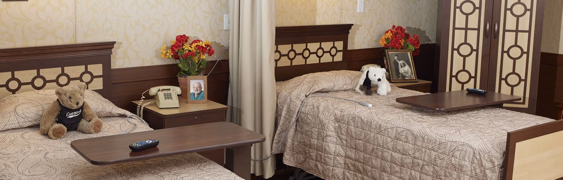 A nursing home room with two single beds separated by a curtain. Each bed has a patterned beige bedspread, a bedside table with a vintage telephone, a framed photo, and a vase of colorful flowers. One bed has a teddy bear wearing a black vest, and the other has a white stuffed dog. Both beds have overbed tables with remote controls on them. The room features wallpaper with a subtle floral pattern and matching wooden headboards and wardrobe doors with geometric designs.