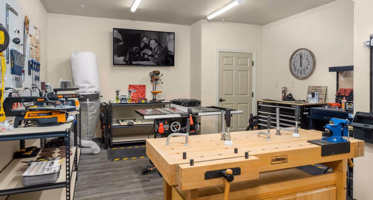 A well-organized workshop room with various woodworking tools and equipment including a large wooden workbench with clamps, a table saw, a drill press, and other power tools. The room has light-colored walls, a clock on the wall, and a black and white photo of two men working on a typewriter mounted above a shelf.