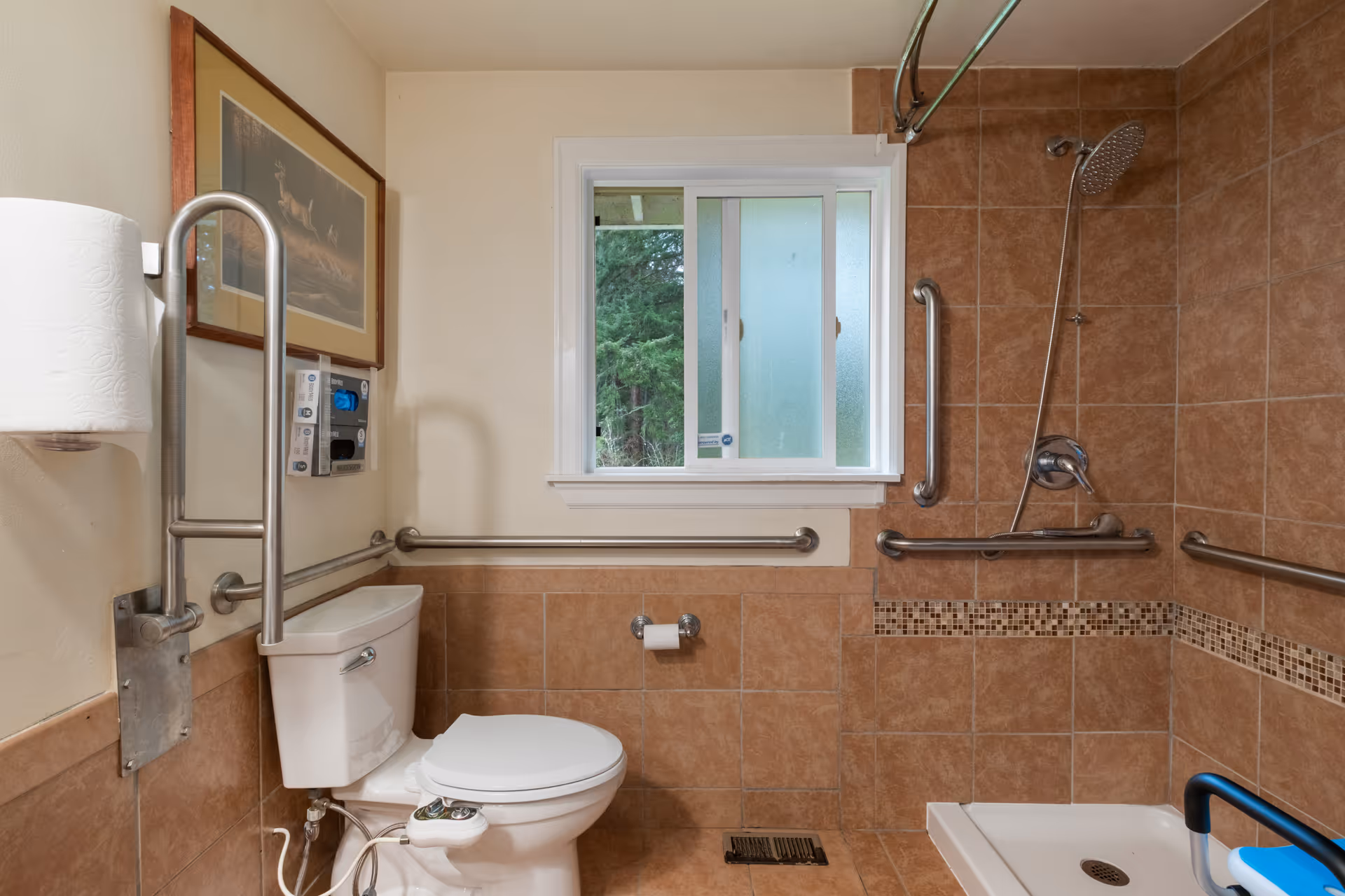 A bathroom with beige tiled walls and floor, featuring a white toilet with support rails around it, a roll of toilet paper on a holder, a framed picture on the wall, a window with frosted and clear glass panes, and a walk-in shower area with multiple grab bars and a handheld showerhead.
