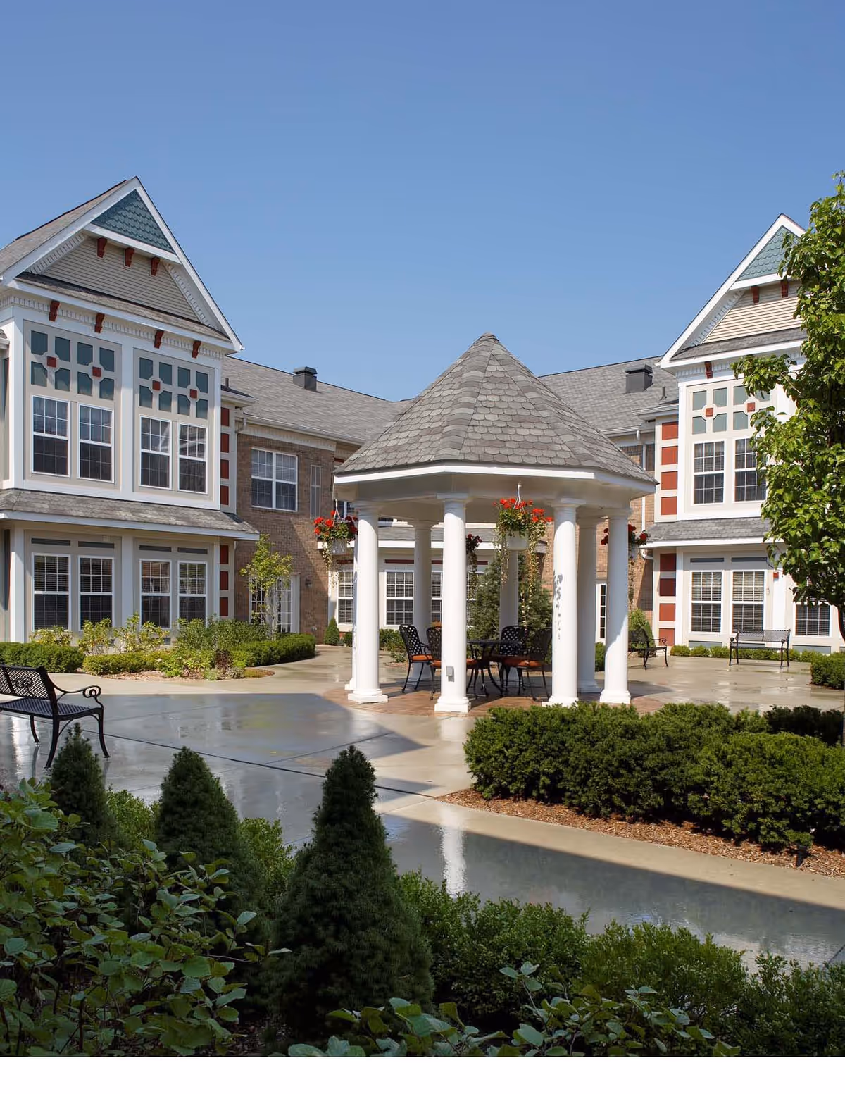 Outdoor courtyard area of a senior living facility with a white gazebo featuring a shingled roof and white columns. Surrounding the gazebo are chairs and tables, landscaped bushes, and a paved walkway. The background shows a multi-story building with large windows and decorative trim under a clear blue sky.