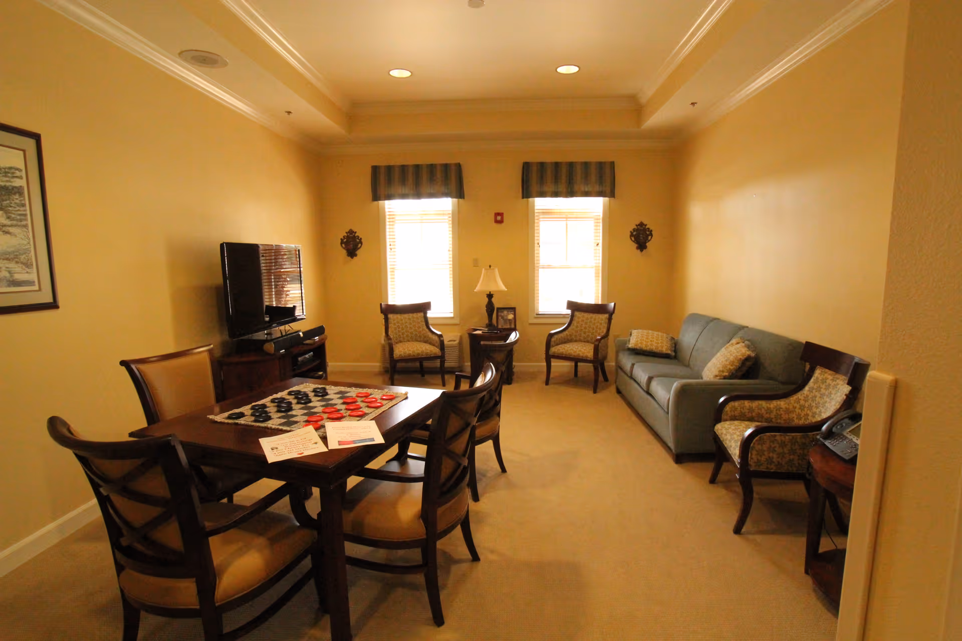 A cozy living room with beige walls and carpet, featuring a wooden table with four chairs and a checkers game set up on it. There is a flat-screen TV on a stand against the left wall, two windows with striped valances, two armchairs, a side table with a lamp, a blue sofa with patterned cushions, and another armchair next to a small table with a telephone.