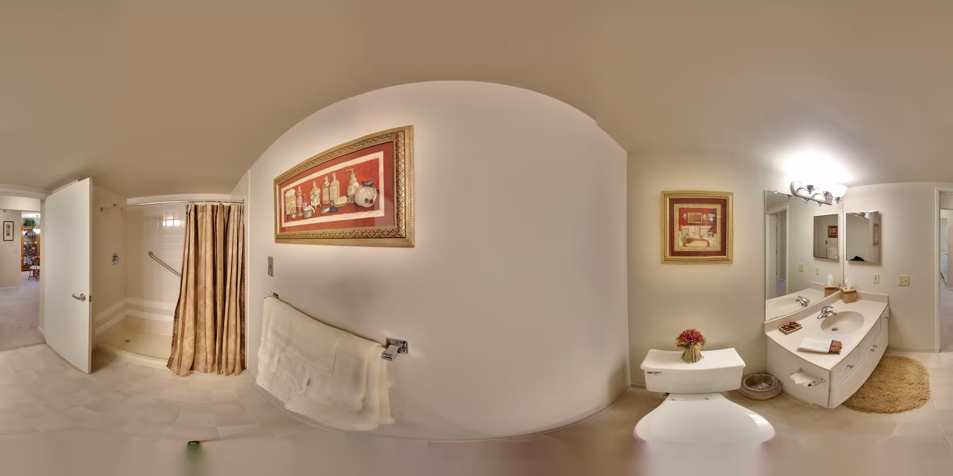 A senior community bathroom featuring a walk-in shower with a beige curtain, a white towel hanging on a rack, a toilet with a small flower vase on top, a vanity with a sink, mirror, and light fixture above, and framed artwork on the walls. The bathroom has light-colored tile flooring and walls, with an open door leading to another room.