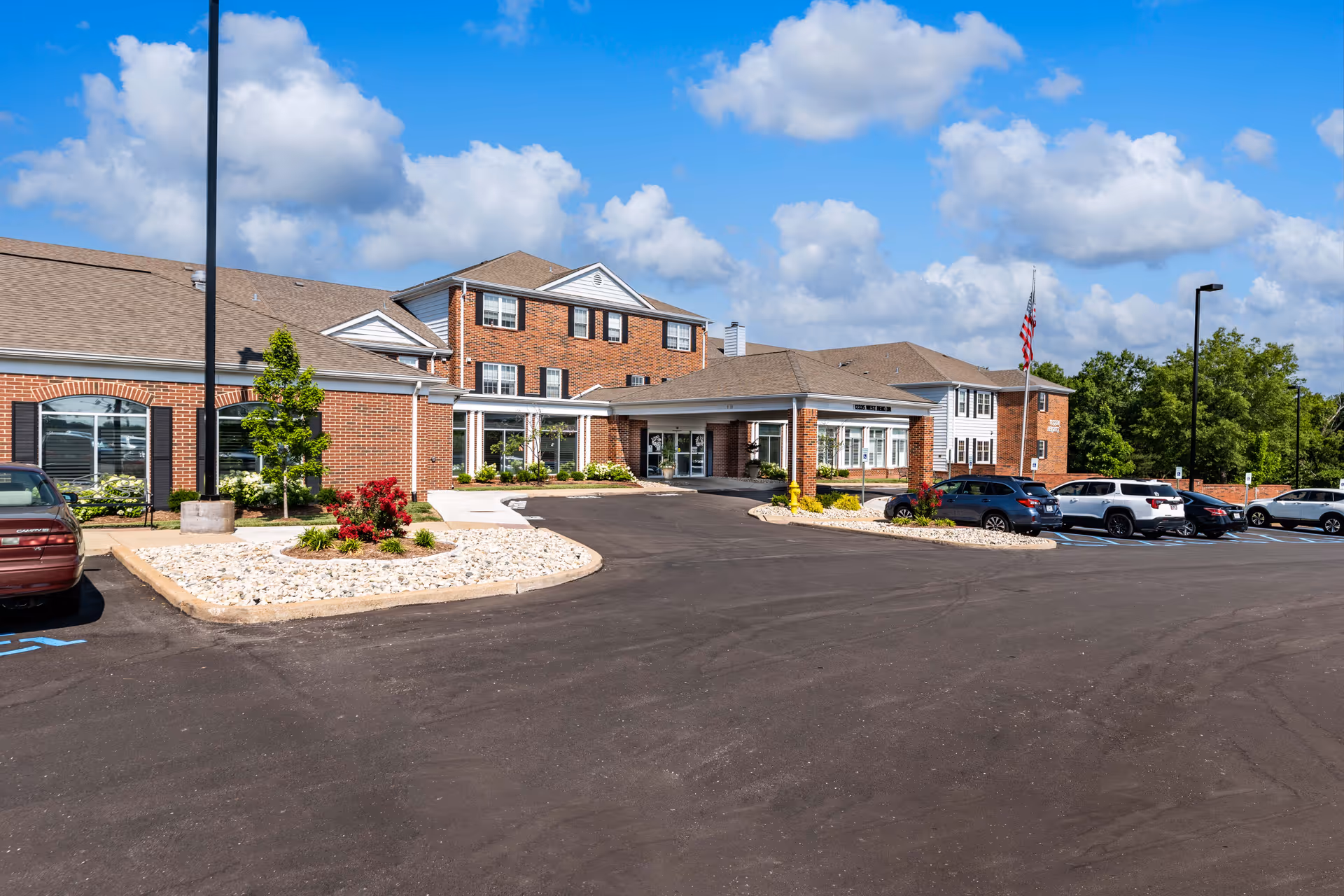Exterior view of Cedarhurst Senior Living of Tesson Heights building with a brick facade, multiple windows, and a covered entrance. Several cars are parked in the parking lot under a partly cloudy blue sky.