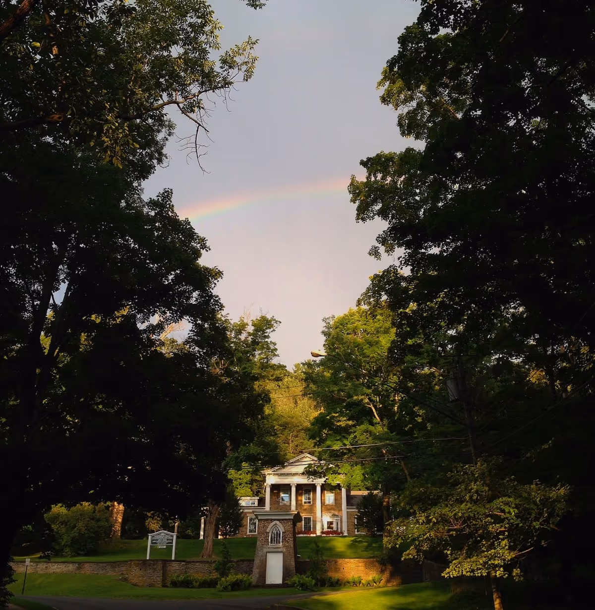 A stately building with white columns partially obscured by large green trees under a sky with a faint rainbow. The building is set on a grassy hill with a stone retaining wall and a small structure with a white door in the foreground.
