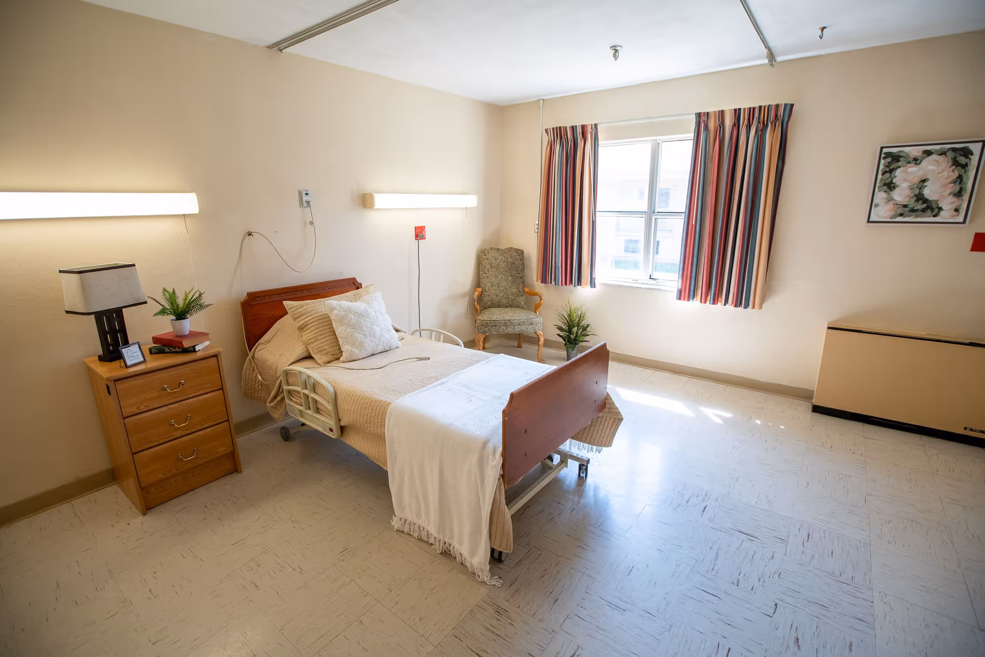 A simple, well-lit nursing care room with a single hospital bed made with beige bedding and a white blanket. Next to the bed is a wooden nightstand with a lamp, a small plant, and some books. There is a cushioned armchair near a window with striped curtains, and a framed floral picture hangs on the wall. The floor is tiled, and the walls are painted a light beige color.