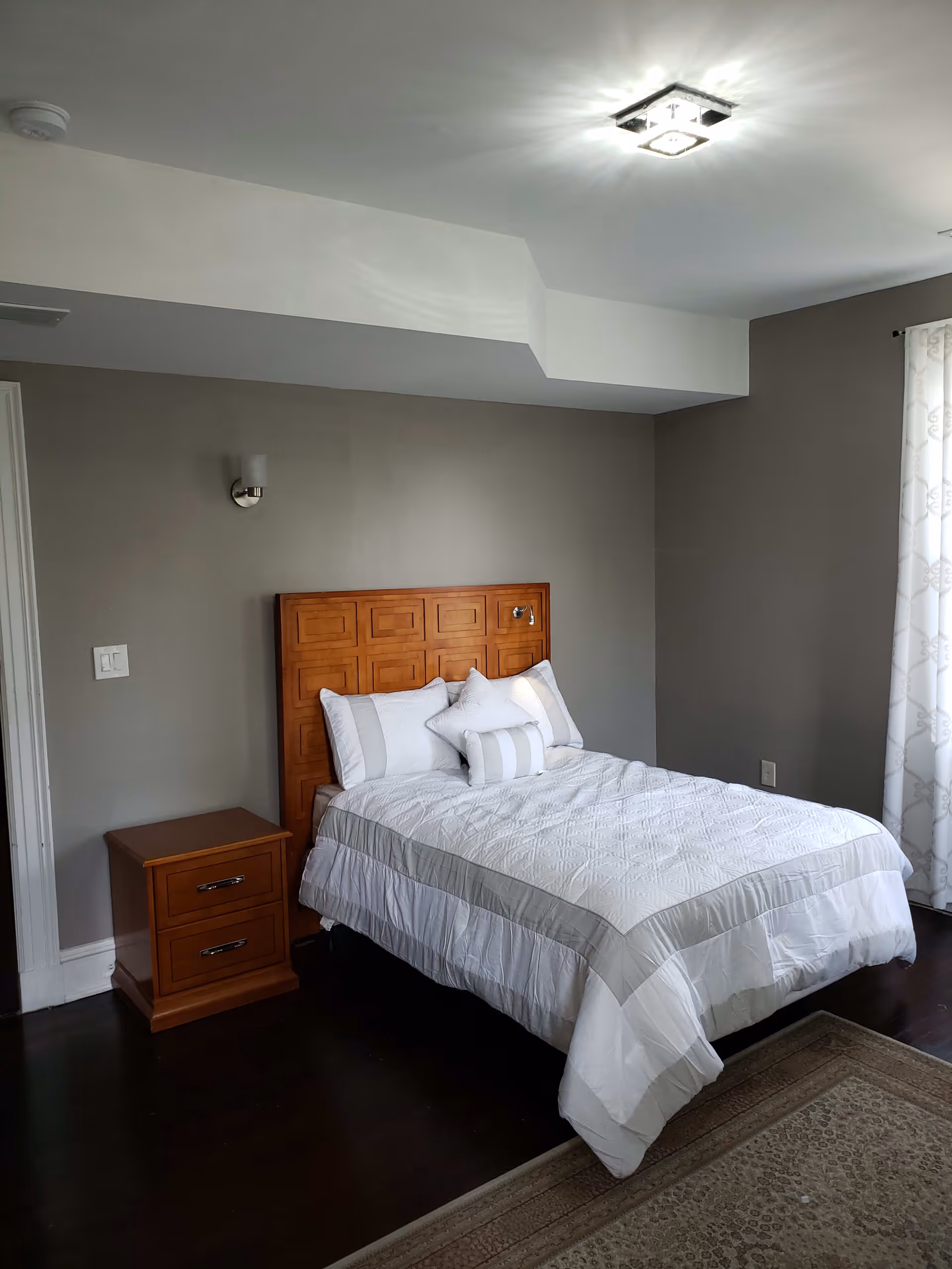 A tidy bedroom with a made bed featuring a wooden headboard, matching nightstand, and gray walls with a window and curtains.