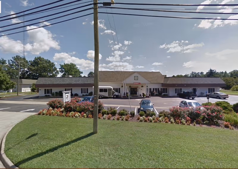 Front exterior view of VIVA Memory Care at Chesapeake building with a parking lot in front, several cars parked, a small bus, and a landscaped area with flowers and bushes under a partly cloudy sky.