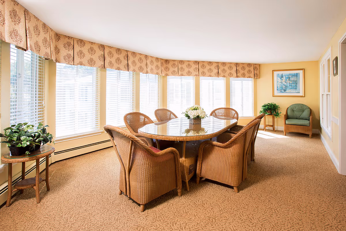A bright and spacious dining room with a large oval glass-top table surrounded by eight wicker chairs. The room features large windows with white blinds and patterned valances, beige carpet, and light yellow walls. There are two small side tables with green plants and a framed painting on the wall.