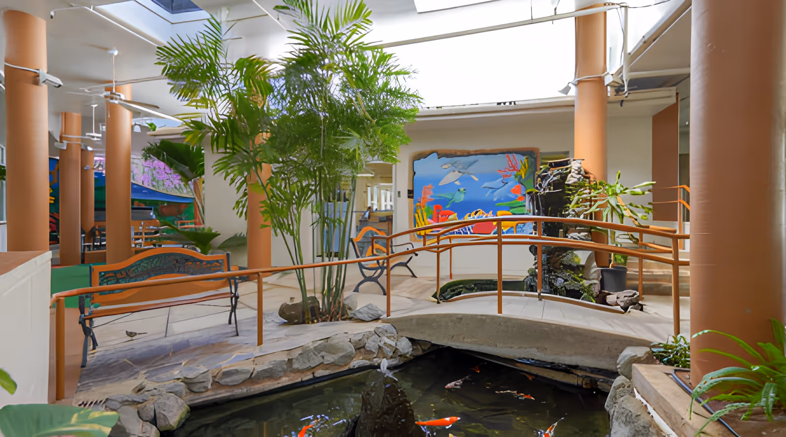 Indoor garden area with a small stone bridge over a koi pond, surrounded by plants and benches. The space has large brown columns and a colorful mural on the wall depicting underwater sea life.