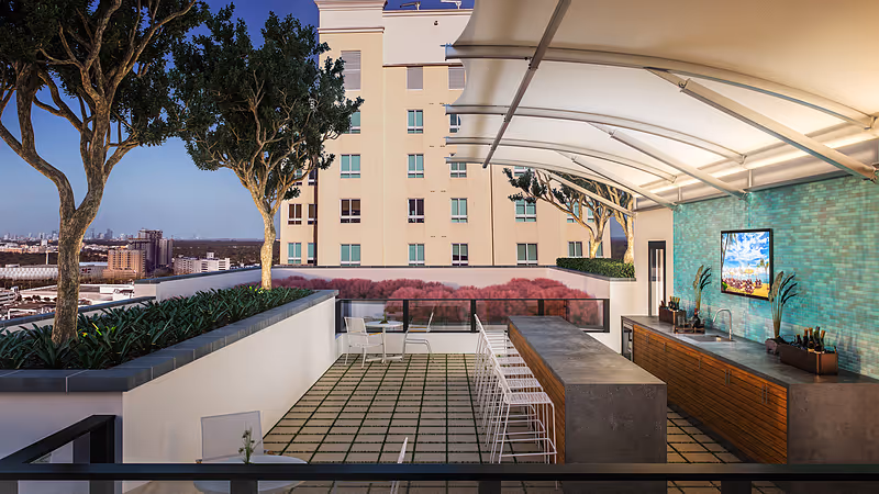 Outdoor rooftop patio area with a modern bar counter and stools, a sink, and a mounted TV on a turquoise tiled wall. There are trees planted along the edge of the patio and a building in the background under a clear sky.